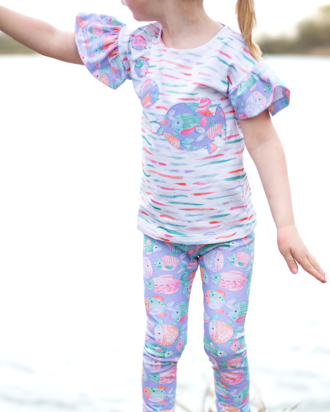 Kinderstoff mit bunten Fischen – Fishtales in Lila. Genähtes Mädchenoutfit: Shirt mit Rüschenärmeln aus Kombistoff mit Fischstreifen, Leggings aus lila Fischstoff. Digitaldruck auf Öko-Tex zertifiziertem Single Jersey.