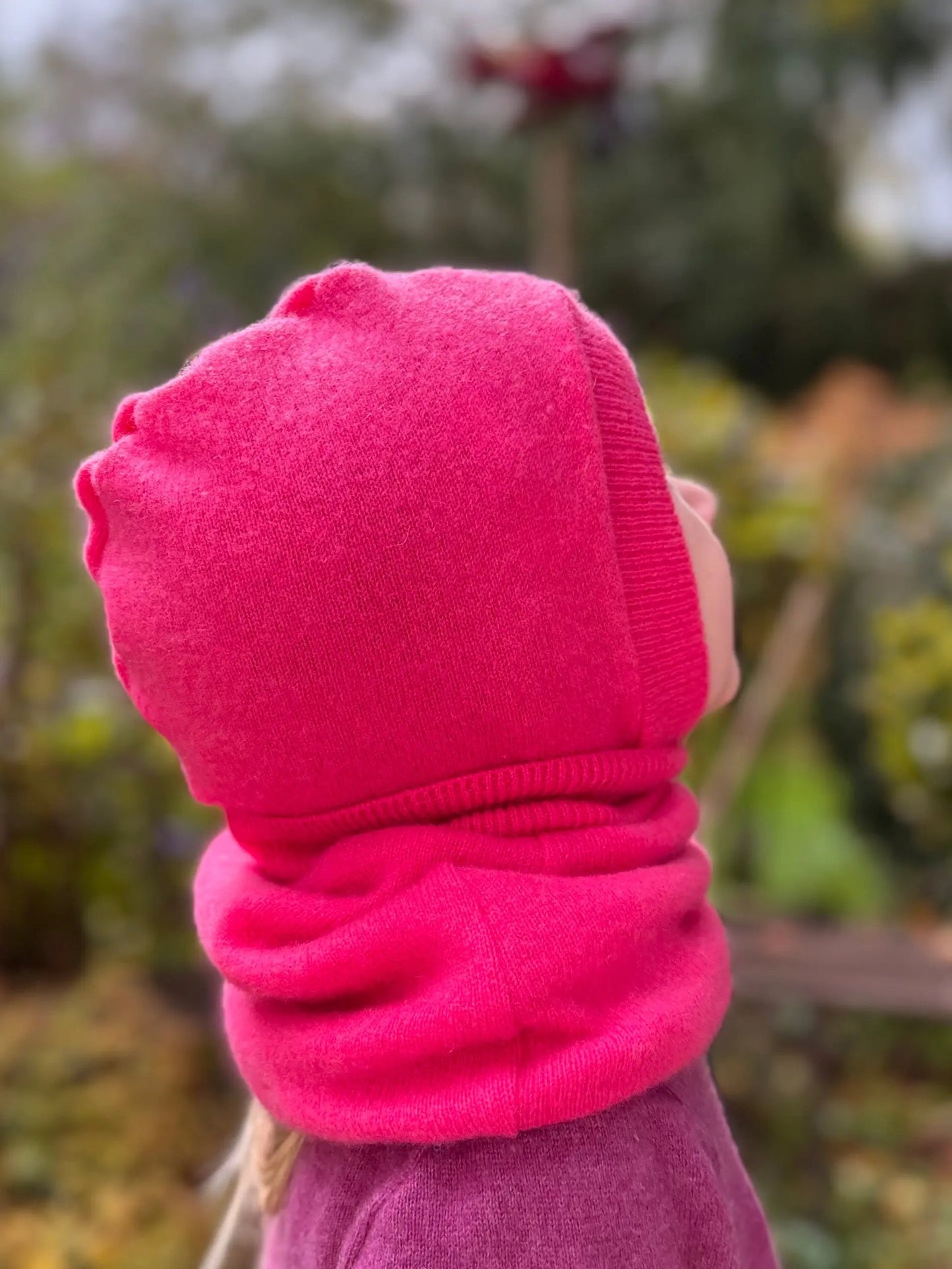 HoodieLooP aus pinkem Jersey, Kind trägt Kapuze und Schal in einem Teil, Rückenansicht im Freien mit Blick auf eine Blüte.