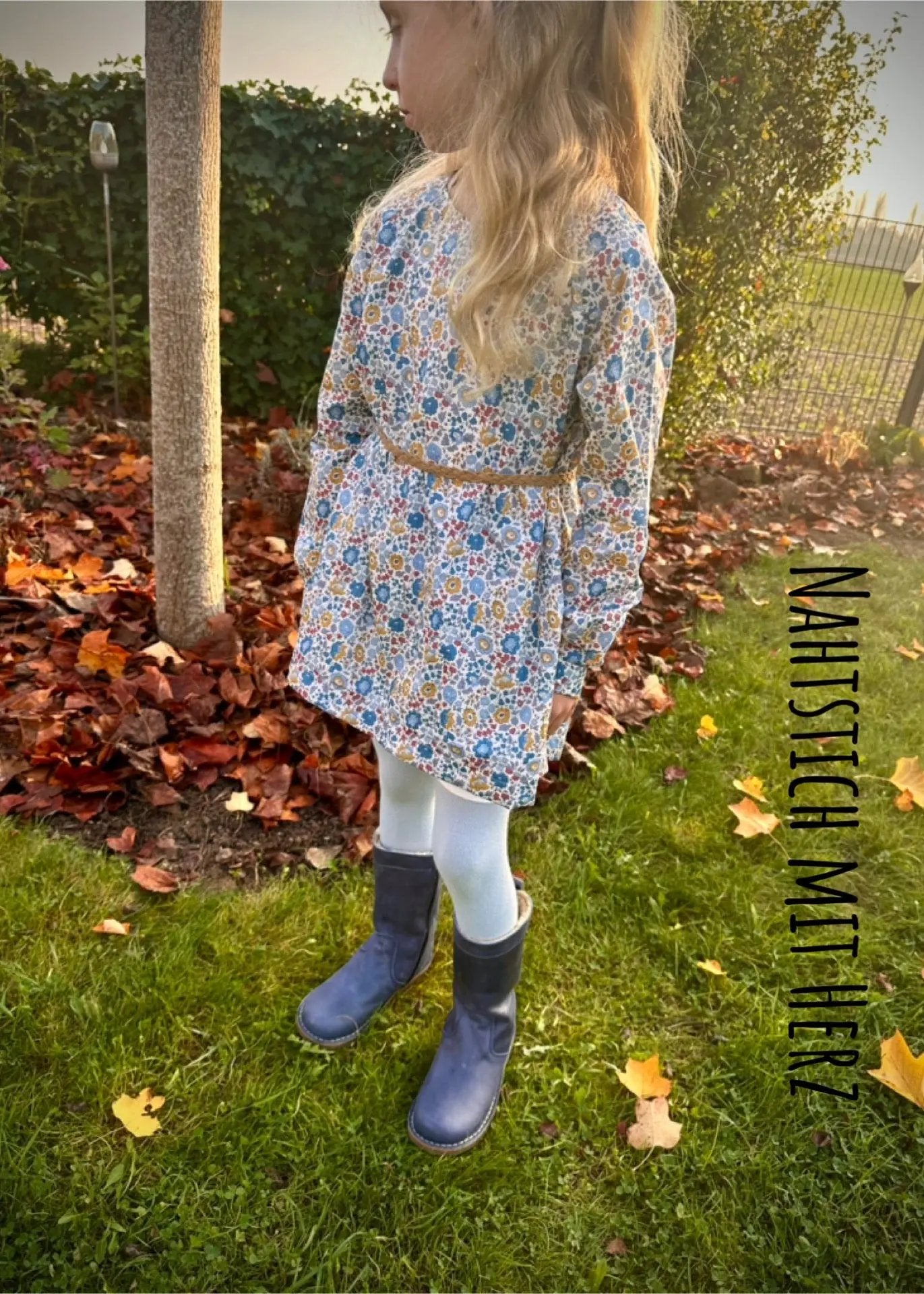 Kinderkleid Ella aus buntem Blumenprint-Musselin mit Kordelgürtel, Kind steht seitlich im herbstlichen Garten.