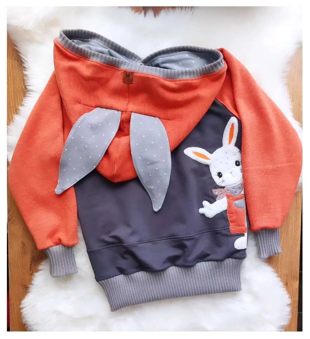 Schnittmuster Hoodie Mika aus orangefarbenem und grauem French Terry – Variante mit langen Hasenohren, Bauchtasche mit Hasen-Applikation, Flatlay auf weißem Fell