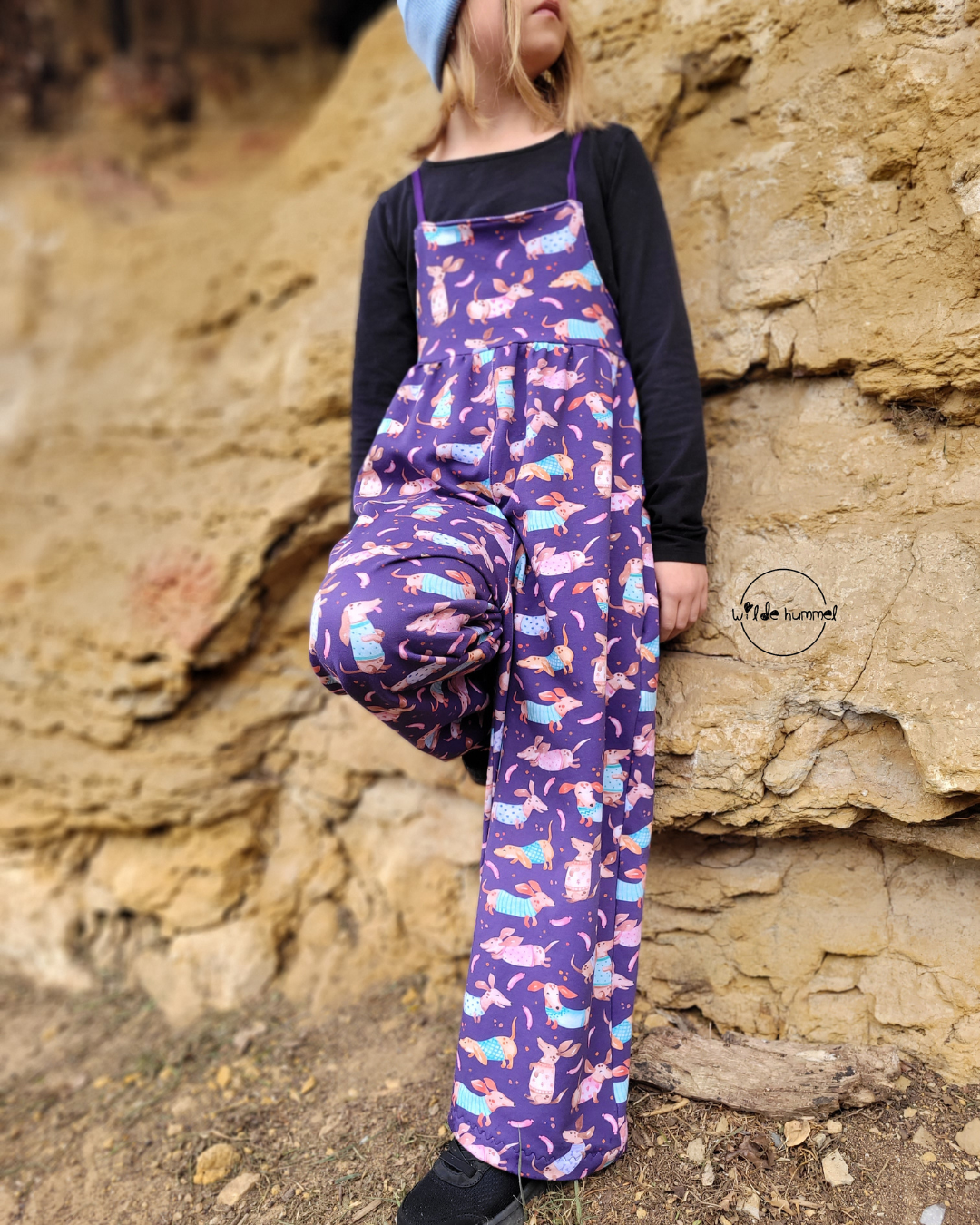 Selbst genähte Kinderlatzhose aus Kinderstoff Dackel Wau Wau Wow Dunkellila – weite Latzhose aus Webware mit Dackeln auf Violett, kombiniert mit schwarzem Longsleeve. Reaktivdruck Meterware, Öko-Tex Standard 100.