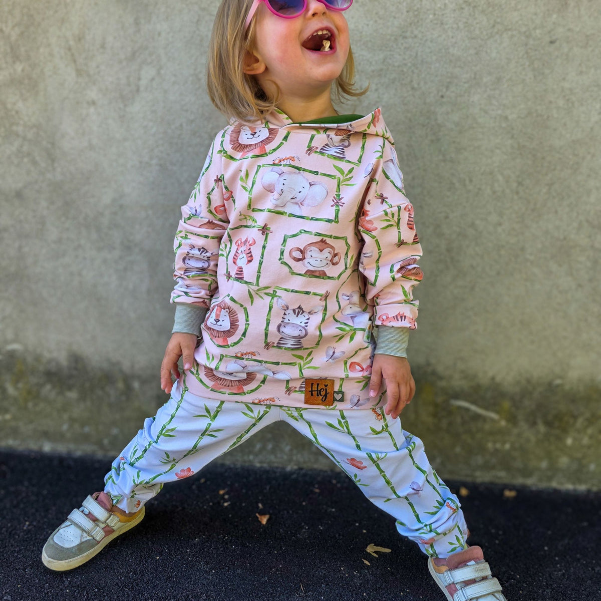 Lachendes Kind in einem Hoodie aus Safari-Tier-Stoff und einer Hose aus Bambus-Kombistoff, beides aus French Terry