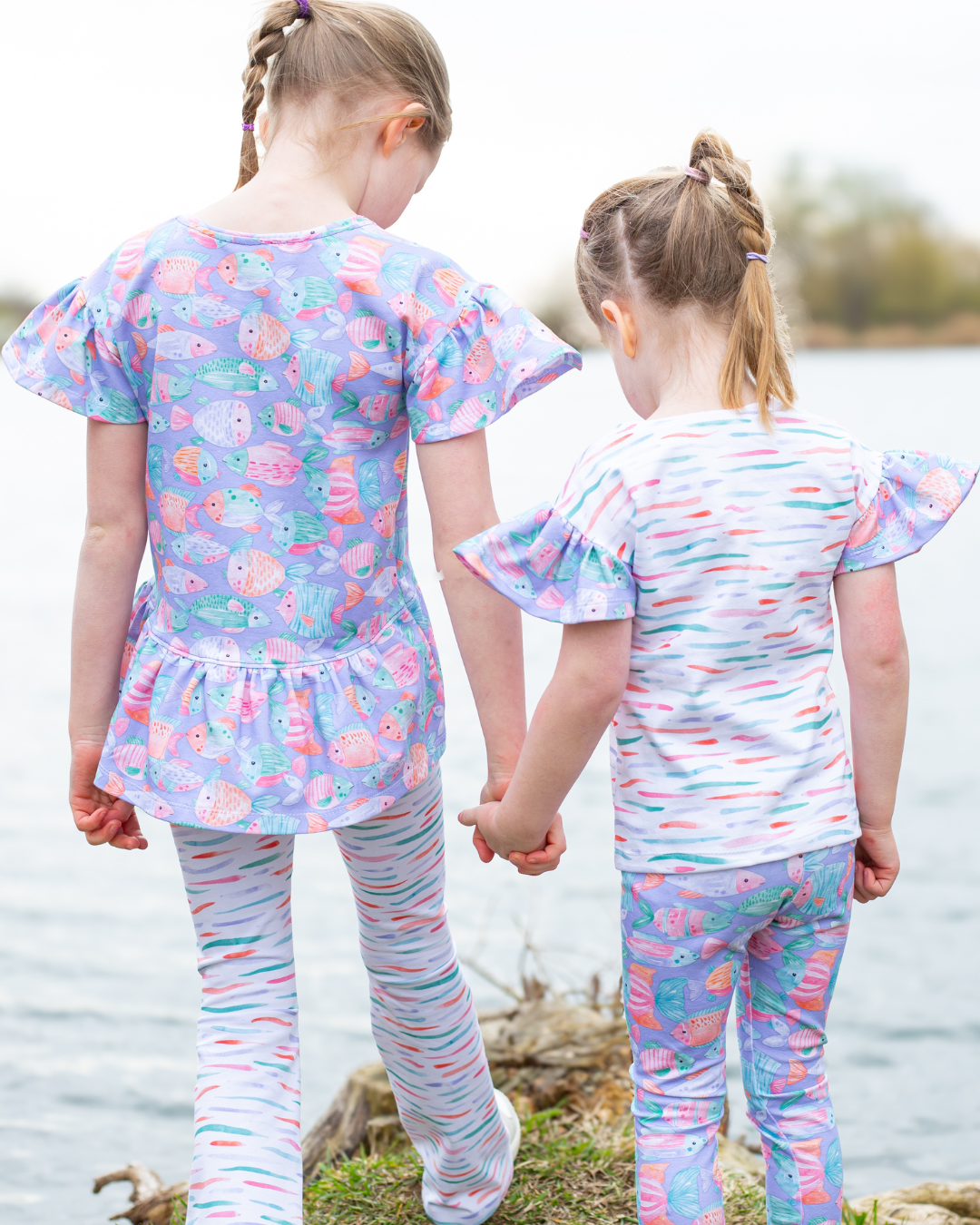 Kinderstoff Fishtales in Lila – zwei Mädchen in selbst genähten Outfits aus der Fishtales-Kollektion. Peplum-Shirt und Leggings aus lila Fischstoff kombiniert mit weißem Kombistoff. Meterware für Kinderkleidung.