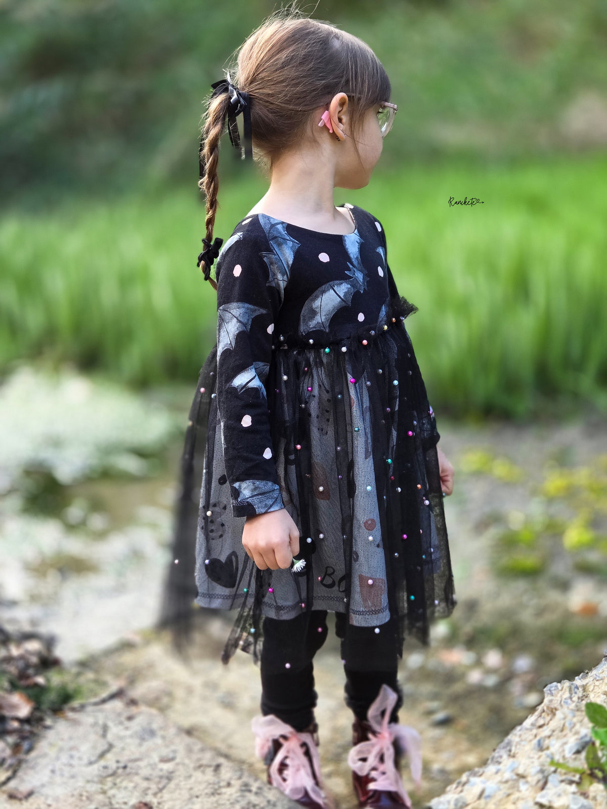 Kind auf einer alten Steintreppe mit Herbstlaub, trägt ein Halloween-Kleid mit Fledermaus-Oberteil und rote Lackstiefel