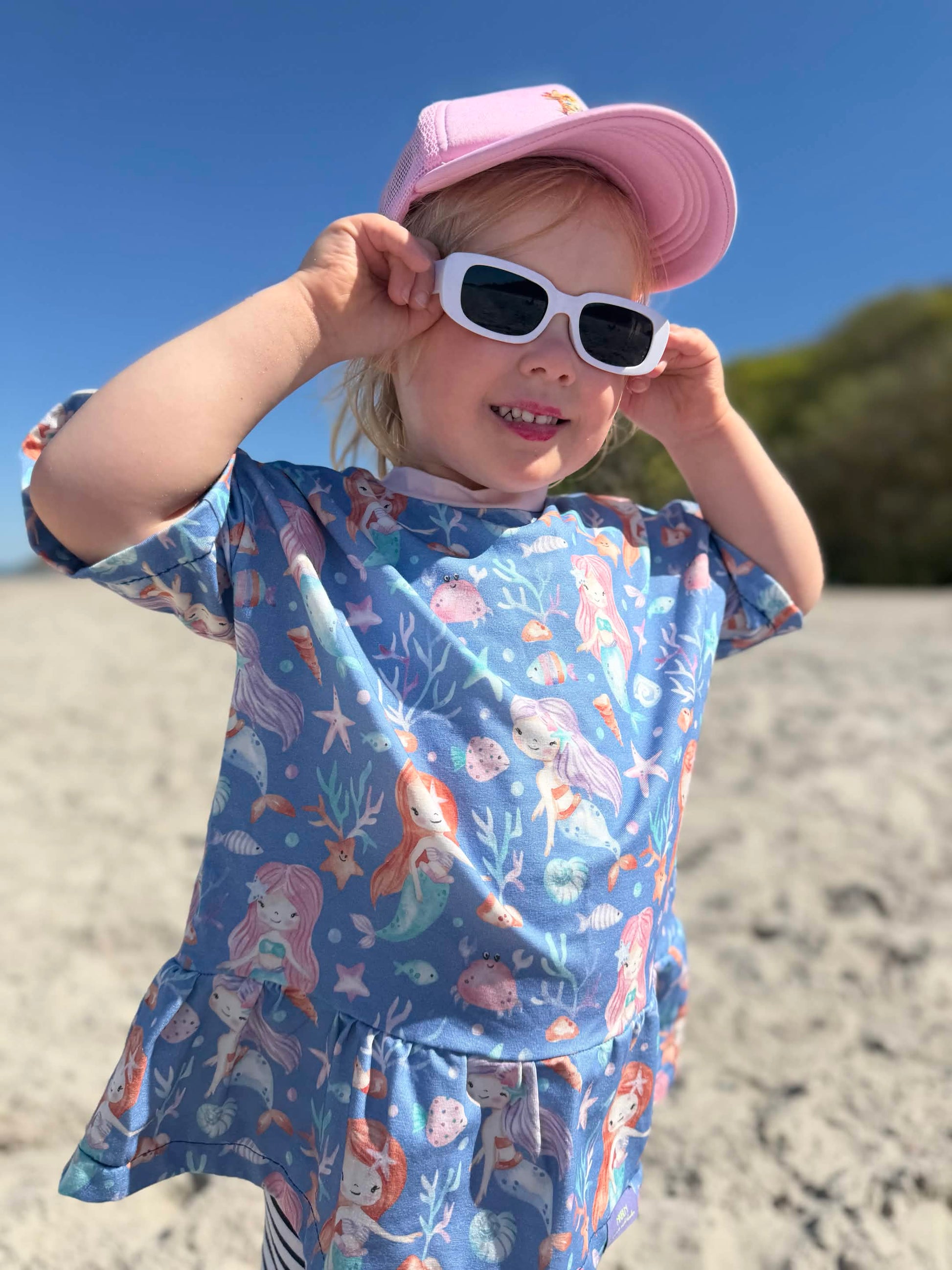 Kind am Strand im selbstgenähten T-Shirt aus Meerjungfrau Stoff in Blau mit handgemalten Meerjungfrauen, Seesternen und Muscheln