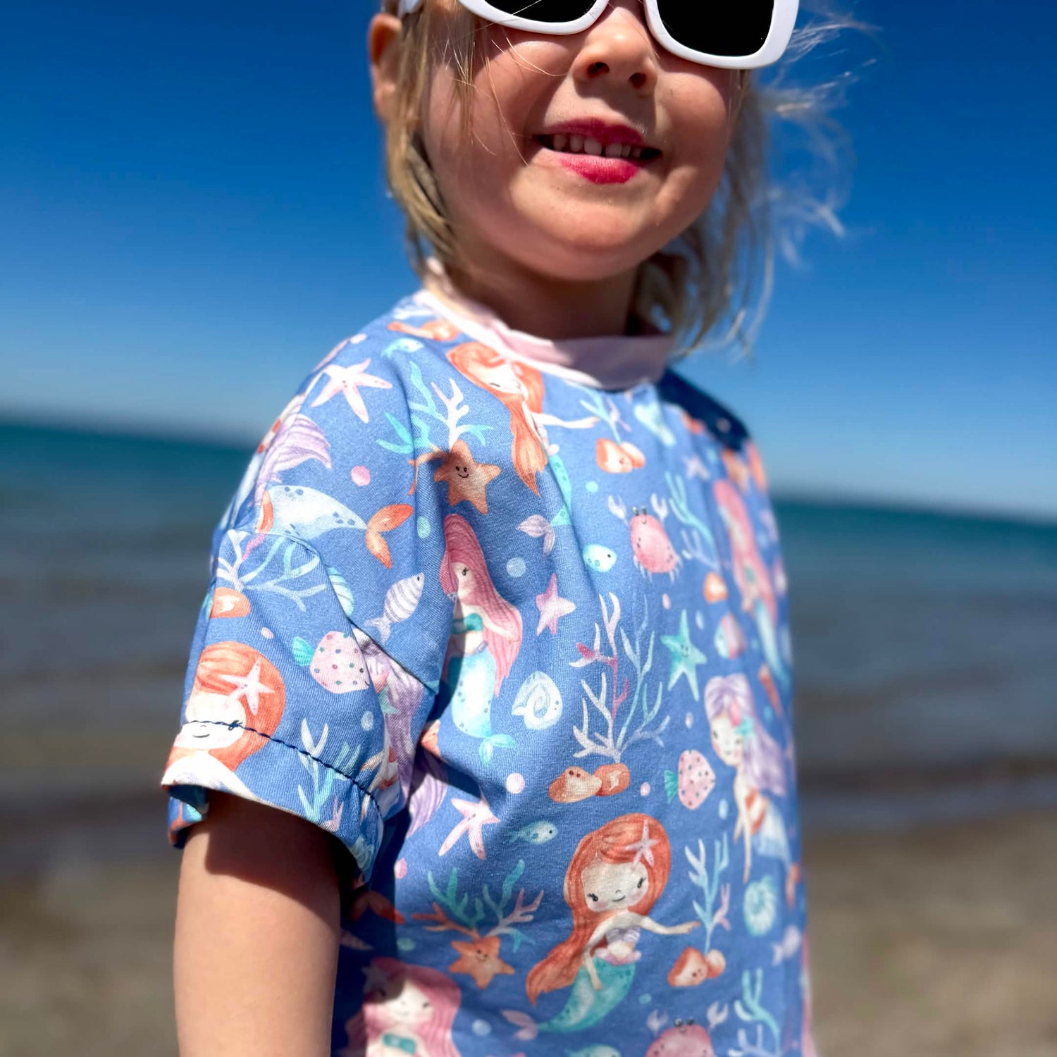 Kinder-Shirt aus Meerjungfrau Stoff in Blau mit handgemalten Meerjungfrauen, Korallen und Seesternen am Meer getragen