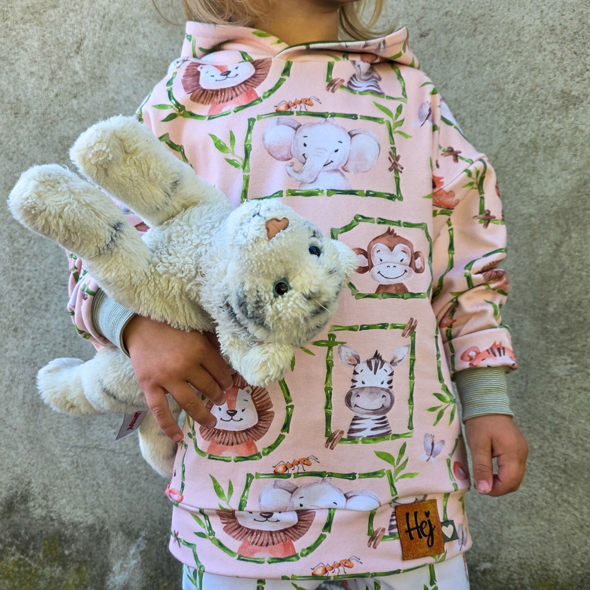 Hoodie aus Kinderstoff Hey Safari mit Elefant, Affe und Zebra in Bambusrahmen, ein Kind hält ein Kuscheltier im Arm