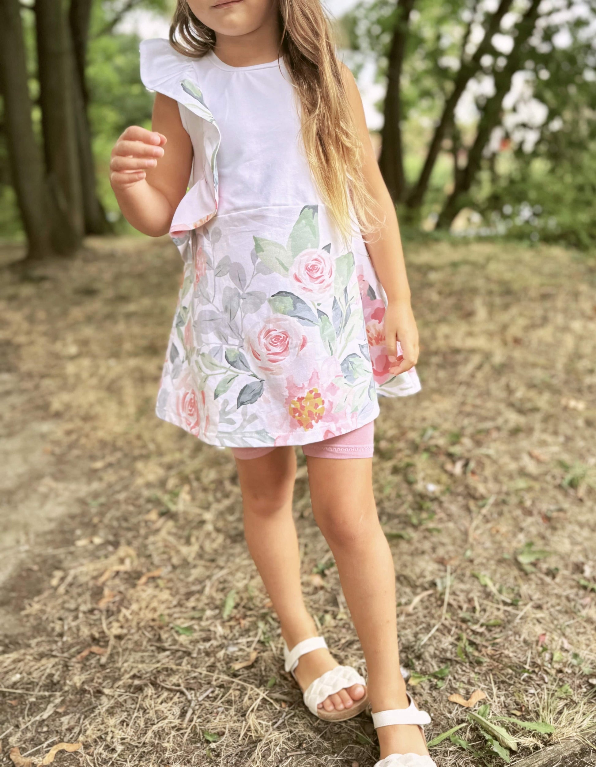 Schnittmuster Kinderkleid Mathilda aus weißer Webware mit Aquarell-Rosen-Bordüre – Variante mit Schulterrüsche, ausgestellter Rock, Kind trägt Kleid im Freien kombiniert mit rosa Radlerhose