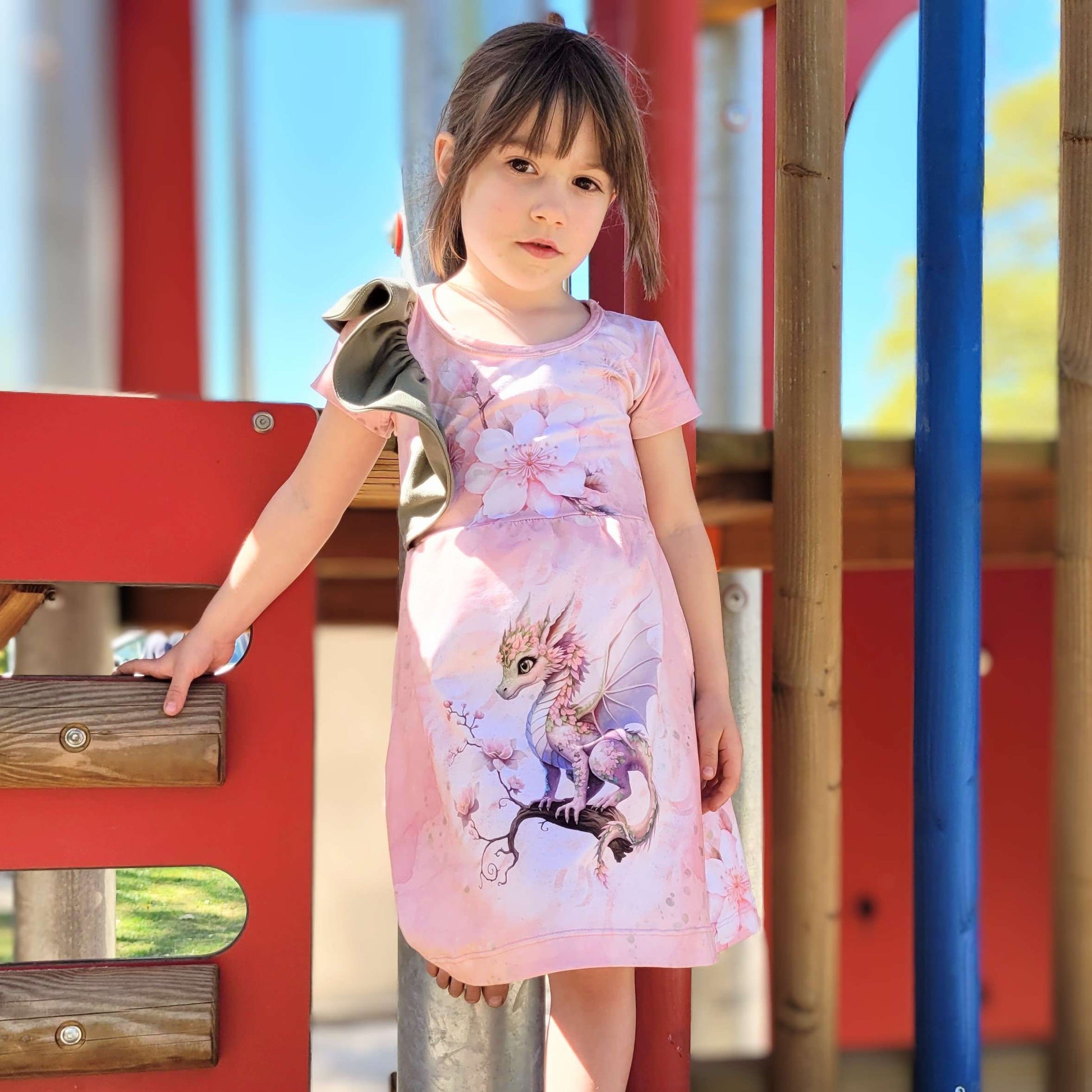 Schnittmuster Kinderkleid Mathilda aus rosafarbenem Jersey mit Drachen-Paneldruck – Variante mit kurzen Ärmeln ohne Rüschen, ausgestellter Rock, Kind auf Spielplatz