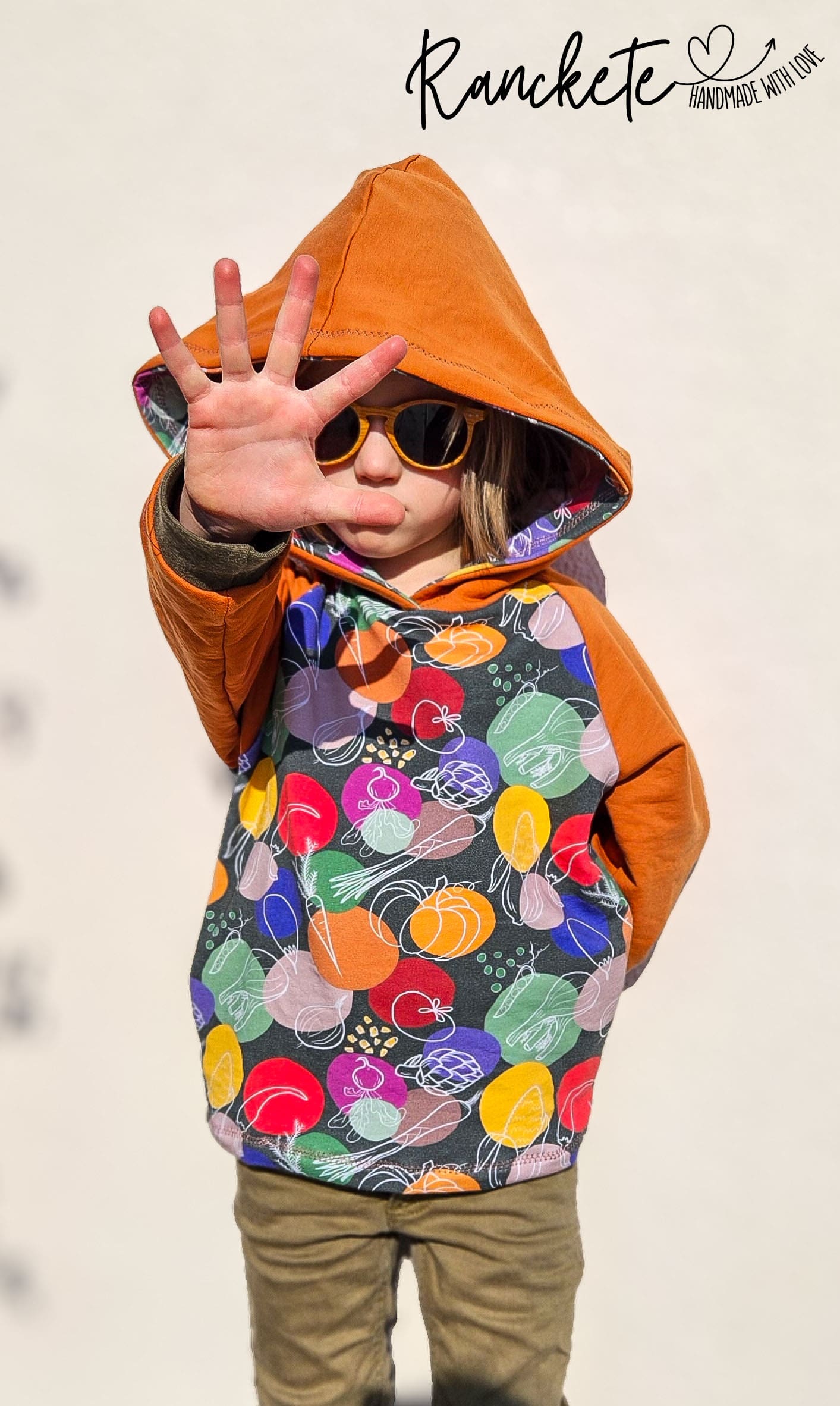 Schnittmuster Hoodie Mika aus dunkelgrauem Jersey mit buntem Gemüseprint – orangefarbene Raglan-Ärmel und Kapuze, Variante ohne Ohren, Kind trägt Hoodie im Freien