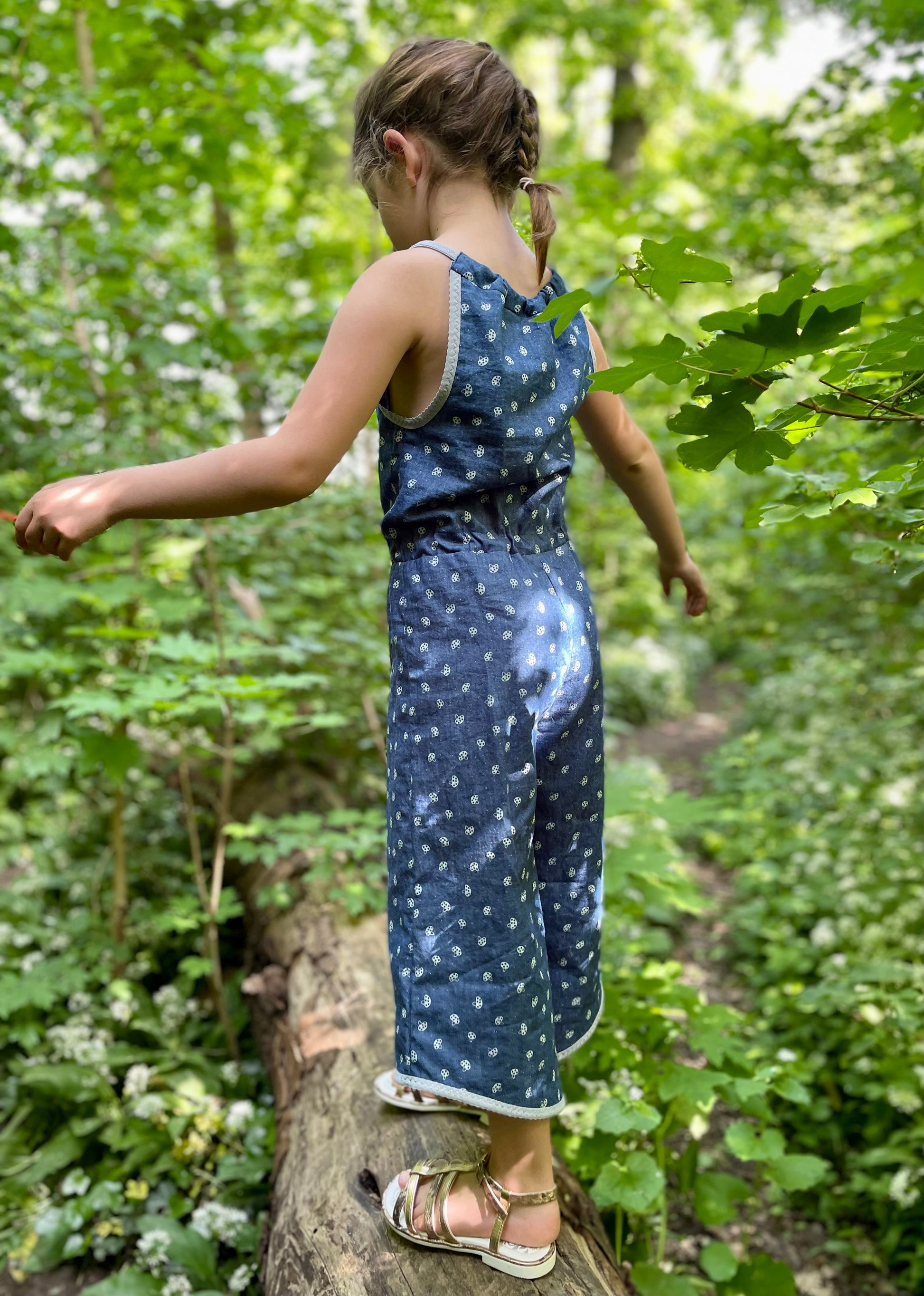 Schnittmuster Jumpsuit Pernille aus blauem Jersey ohne Rüschen – Vorderansicht mit sichtbarem Kordelzug und Taillenbund, Kind auf Baumstamm im Wald, lange Hosenvariante