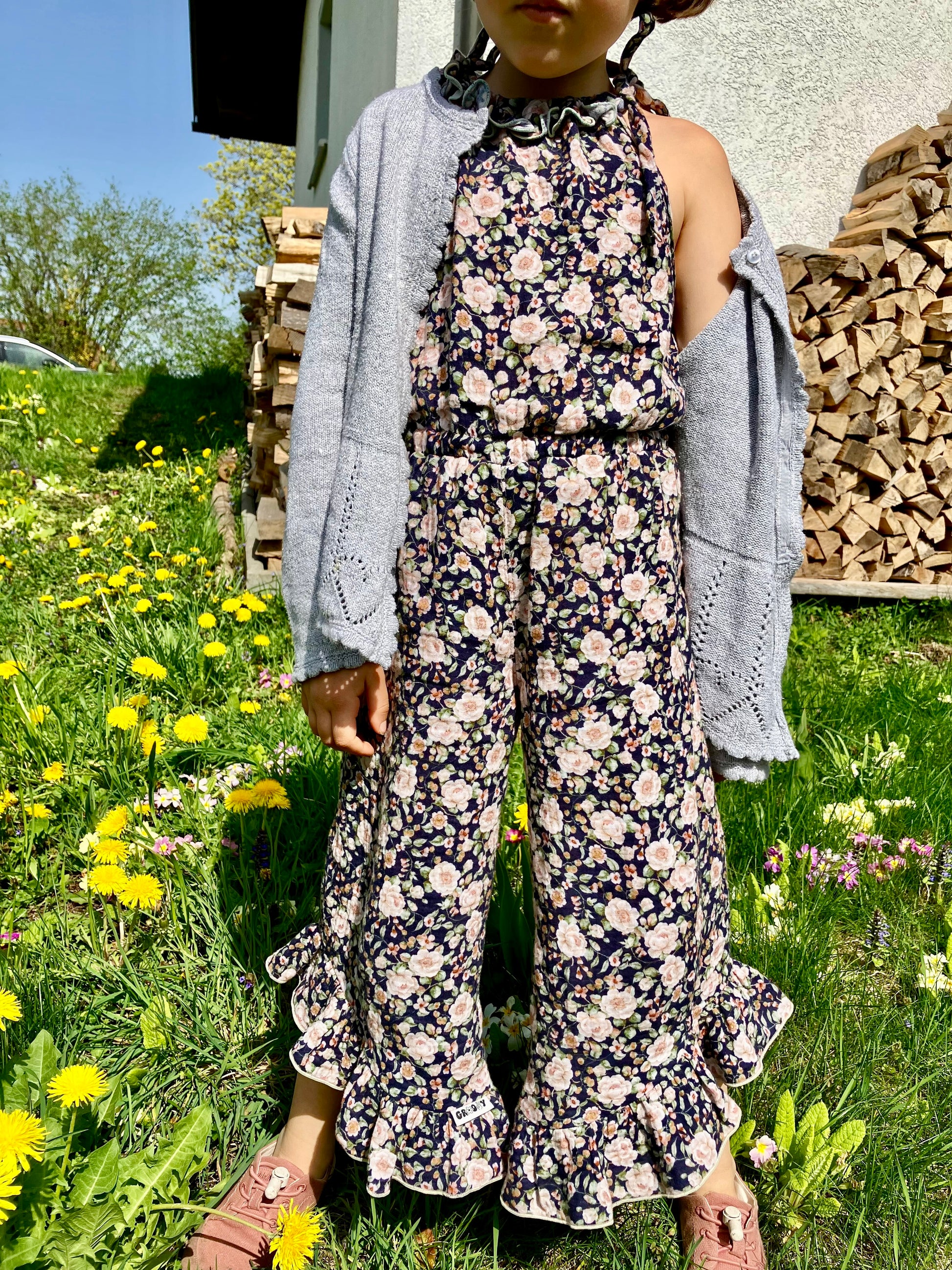 Schnittmuster Jumpsuit Pernille aus dunkelblauem Webware mit Rosenprint – lange Hosenvariante mit Rüschen am Saum, kombiniert mit grauer Strickjacke, Kind im Freien