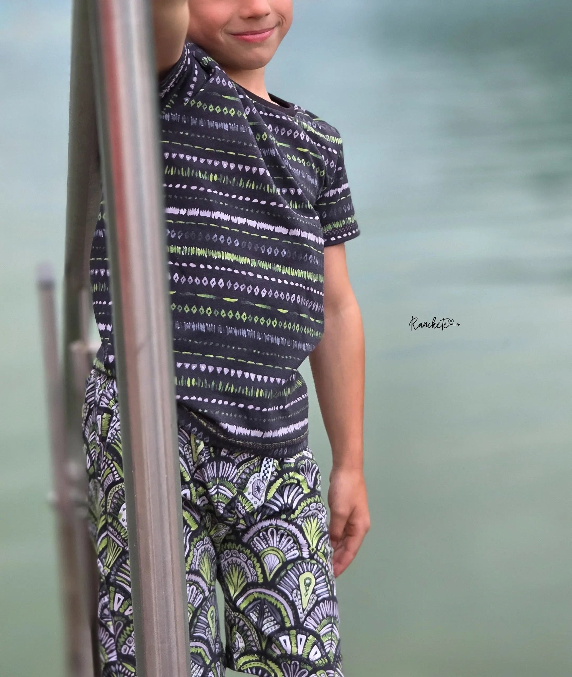 Kind trägt Shirt aus Streifenstoff Grün Schwarz kombiniert mit Shorts aus dem Ornamentstoff Emerald Reverie.