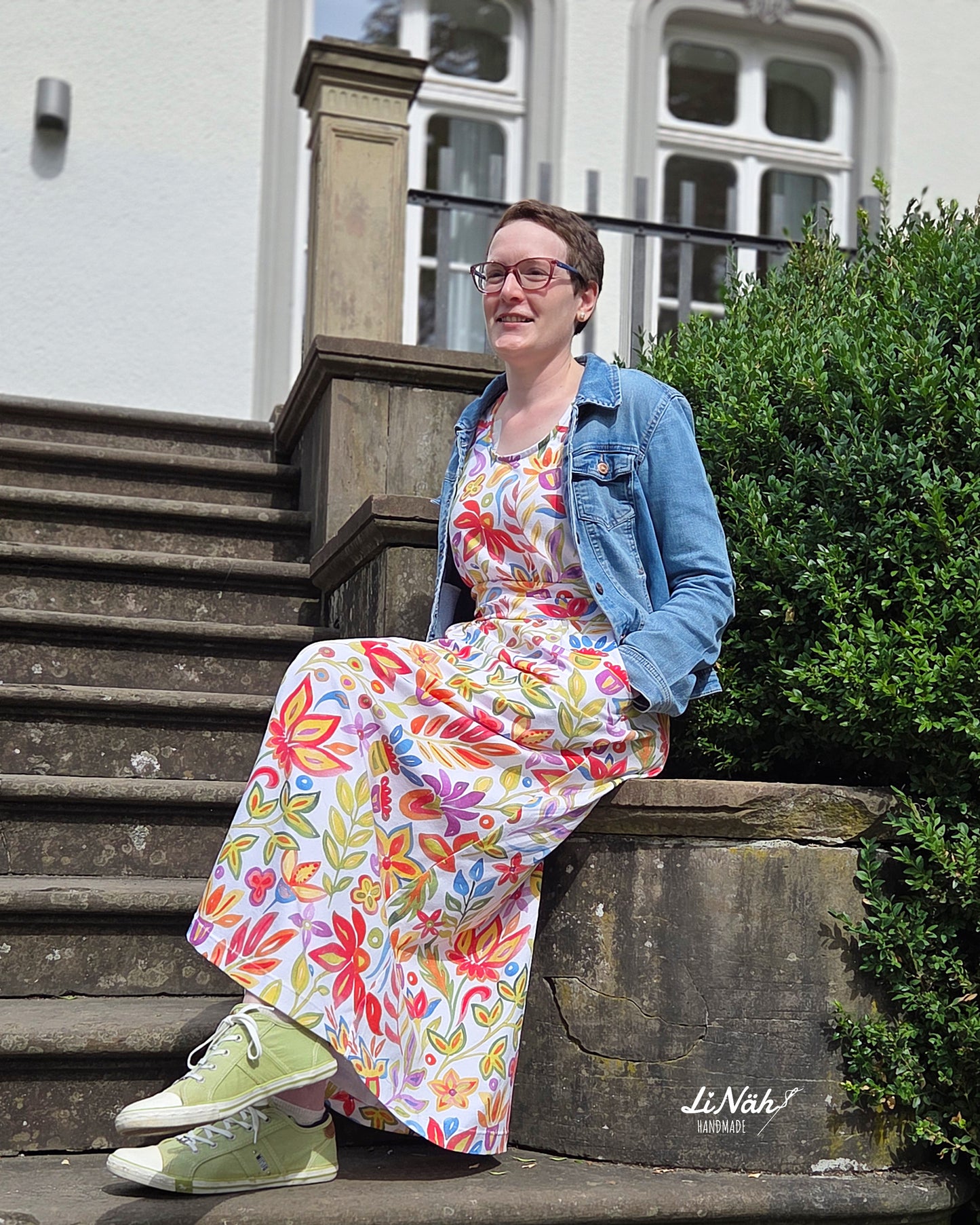 Frau in Farbenfest Maxikleid und Jeansjacke sitzt lächelnd auf einer Steintreppe vor einem historischen Gebäude
