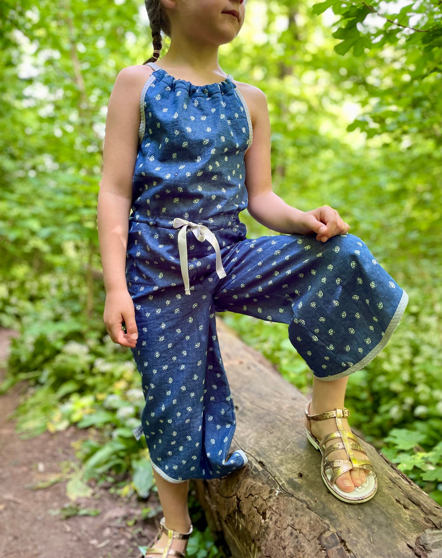Schnittmuster Jumpsuit Pernille aus blauem Jersey mit Punktemuster – lange Hosenvariante, Rückenansicht, Kind balanciert auf Baumstamm im Wald, Gr. 98–110