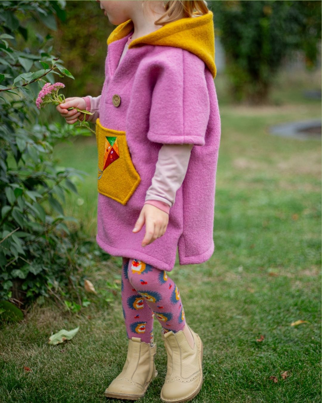 Schnittmuster Poncho Jonte aus rosafarbenem Wollwalk – Variante mit Kapuze und Bauchtasche aus Kontrastfarbe mit Drachen-Applikation, Kind im Garten im Herbs