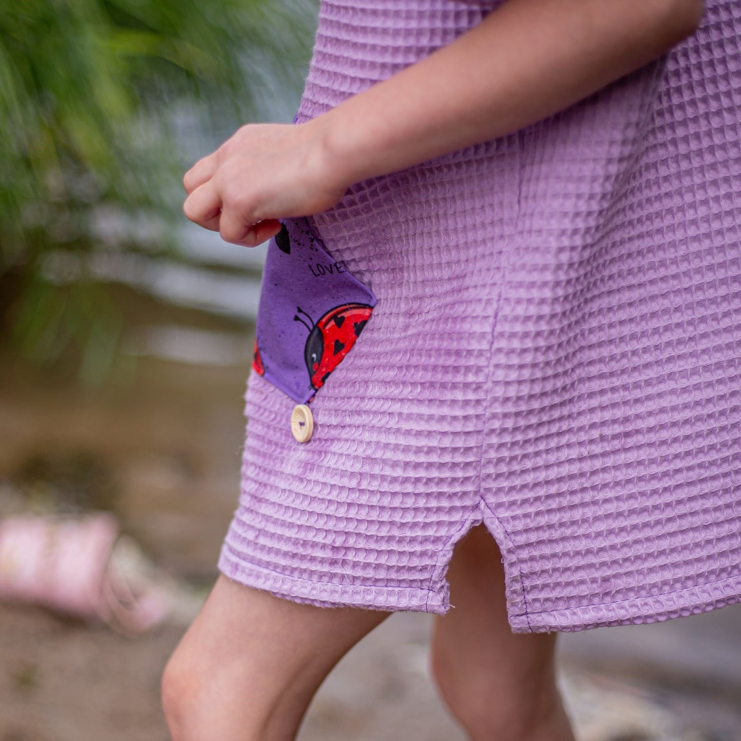 Schnittmuster Poncho Jonte aus lila Waffelpiqué – Badeponcho mit Kapuze und Bauchtasche aus Marienkäfer-Print, Kind steht barfuß am Wasser