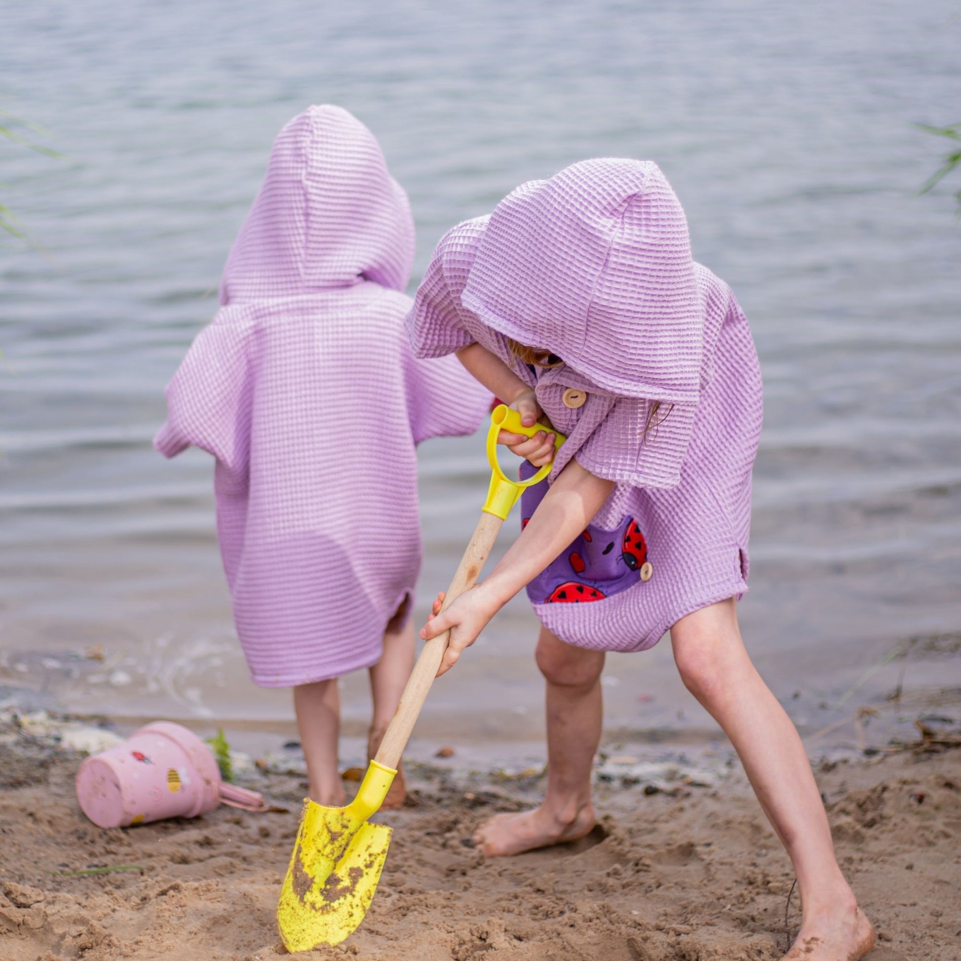 Schnittmuster Poncho Jonte als Badeponcho aus lila Waffelpiqué – zwei Kinder spielen am Sandstrand, Variante mit Kapuze, Gr. 98–110