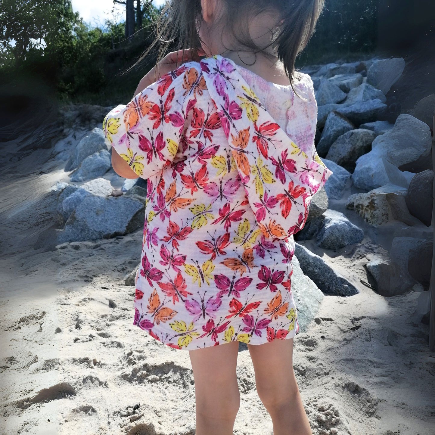 Schnittmuster Poncho Jonte aus weißem Musselin mit buntem Schmetterlings-Print – Variante ohne Kapuze als leichter Sommerponcho, Kind steht barfuß am Sandstrand